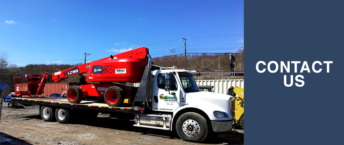 An LGMG 65' boom lift is loaded up on the back of a trailer. The tractor trailer is in a parking lot with a lot of equipment and blue sky overhead. 